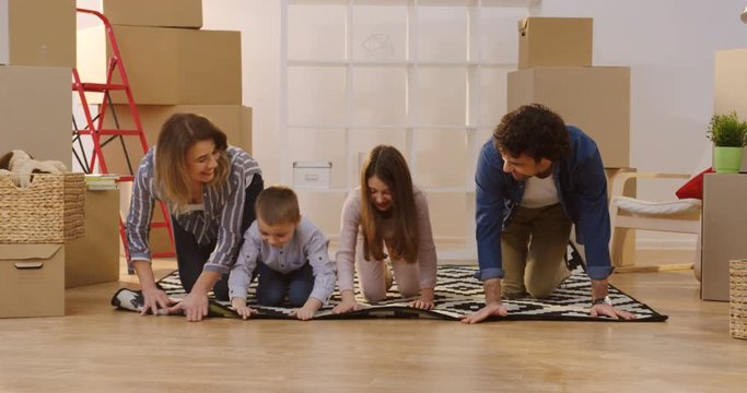 Cheerful Caucasian family unpacking the patterned carpet in the cozy room full of the boxes while they moving in the new flat. Inside