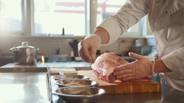 Butcher adding spices into large piece of fresh raw meat lying on a wooden board in a commercial kitchen of restaurant