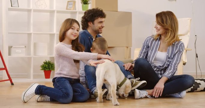 Portrait shot of the happy Caucasian family with a labrador puppy sitting on the floor surrounded by the many boxes and home stuff during moving in the new houses. Indoor