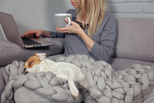 Cozy home, woman covered with warm blanket drinking coffee, Dog sleeping on female feet. Relax, carefree, comfort lifestyle.