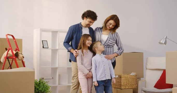 Portrait shot of the happy parents with kids standing in the cozy living room full of unpacked boxes and posing with keys from a new house. Indoors
