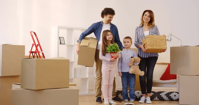 Portrait of the cheerful Caucasian family with kids standing in the nice living room full of unpacked boxes and smiling to the camera with boxes and stuff in hands while moving in the new house