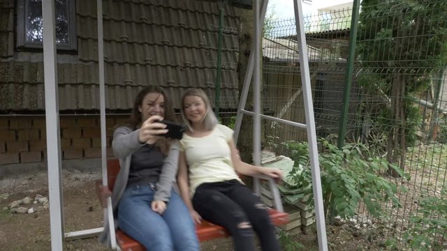 Couple of pretty teenage female friends taking a selfie with smartphone while they swinging outdoor relaxing together in the cradle 