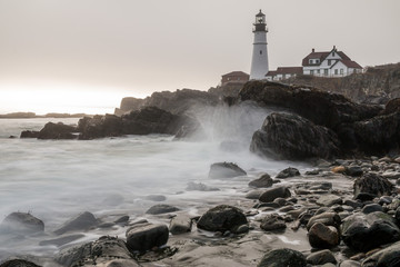 Fototapeta na wymiar A foggy, rocky beach in Maine with Portland Head Lighthouse in the background. 