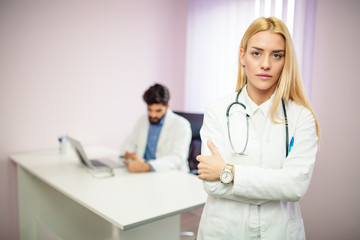 Portrait of blonde serious female doctor while standing in front of camera, male doctor in background working on the computer.