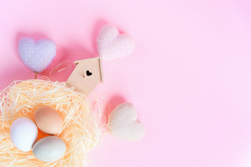 Easter eggs of different colors in a straw nest, wooden bird feeder and decorative textile hearts on a pink background,Top view free copy space. Flat lay. Easter concept.
