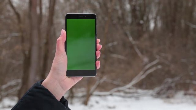 Mockup video of black mobile telephone in female hand. Front view of modern cellphone with blank green screen isolated at blurry snowy winter background in park or wood.