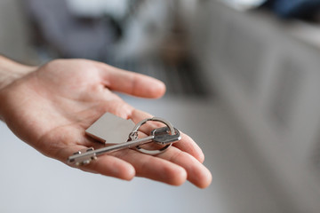 Men hand holding key with house shaped keychain. Modern light lobby interior. Mortgage concept. Real estate, moving home or renting property.