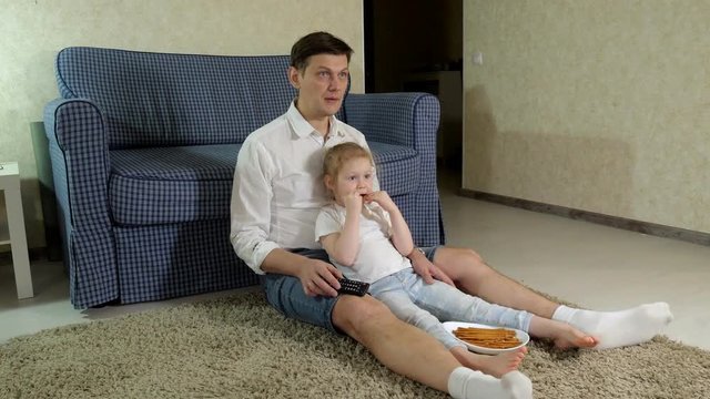 man and daughter watching television, sitting on the floor eating snacks