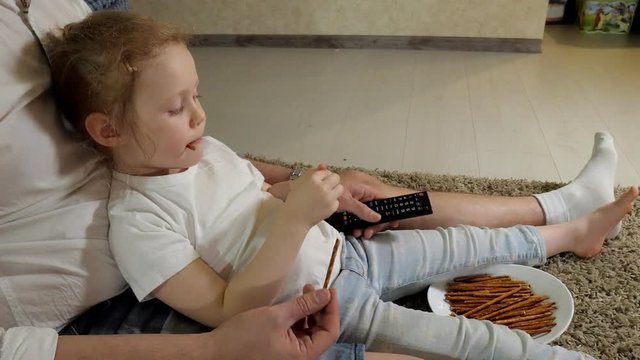 man and daughter watching television, sitting on the floor eating snacks