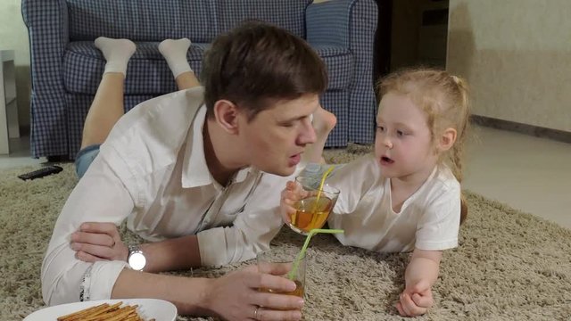 man and daughter watching television, sitting on the floor drink juice