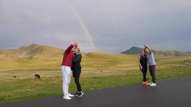 Romantic couples in love enjoying countryside trip taking selfies with smartphone hugging and kissing with idyllic hillside and amazing rainbow in background