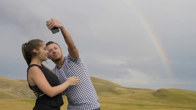 Passionate couple of teen lovers with smartphone taking selfies pics and photographing the picturesque countryside panorama rainbow