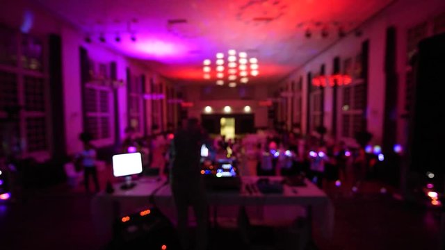 Defocused rear view of music DJ mixing dancing in front his turntables and mixer and laptop during party wedding - illumination laser light of his back large room macarena dancing guests