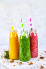 Green, yellow, purple smoothies in bottles of berries, greens, oranges on a white board on a gray table.