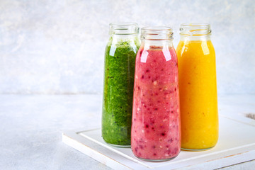 Green, yellow, purple smoothies in bottles of berries, greens, oranges on a white board on a gray table.