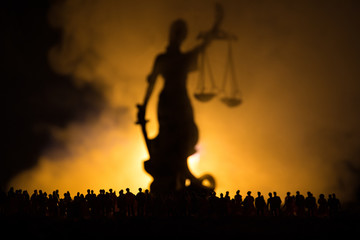 Fototapeta premium Silhouette of blurred giant lady justice statue with sword and scale standing behind crowd at night with foggy fire background. at night