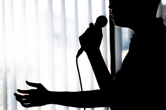silhouette Young man Singer is singing or speaking in Concert raising his hands with microphones on light event convention hall light background