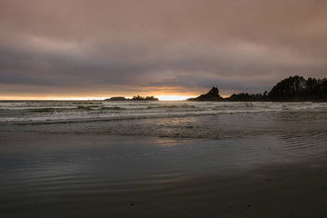 A distant view of a cloudy sunset as the ocean waves glide across the sandy beach and a silhouette of Pettinger Point. 