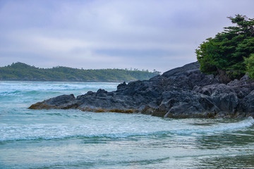 A cloudy afternoon as ocean waves crash against rocks and there are trees in the distance.