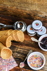 A Kit For Making Ice Cream: Spoon For Ice Cream, Ice Cream Cones, Jam, Topping, Chocolate, Sprinkles, Coconut Shavings. On Wooden Background. The Concept Of A Set, Surprise, Gift Idea