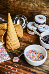 A Kit For Making Ice Cream: Spoon For Ice Cream, Ice Cream Cones, Jam, Topping, Chocolate, Sprinkles, Coconut Shavings. On Wooden Background. The Concept Of A Set, Surprise, Gift Idea