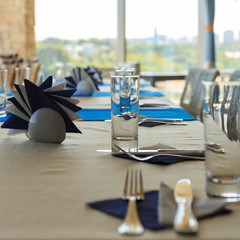 Empty half-served banquet table in restaurant with napkins, glasses, forks, knives, shallow DOF view against blurred city skyline