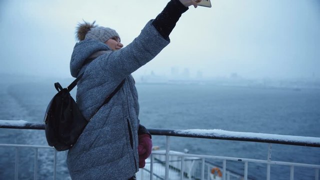 Casual woman in warm winter clothes and with backpack standing on frost ship deck and taking selfie with smartphone on gloomy seascape.