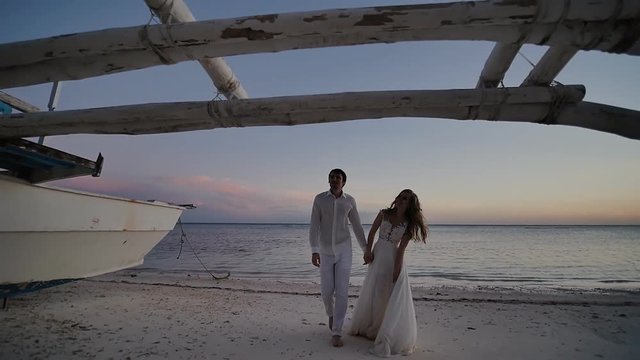 The bride and groom, newlyweds, walk at sunset on a tropical beach by the ocean. They are posing against the backdrop of an old Philippine boat.