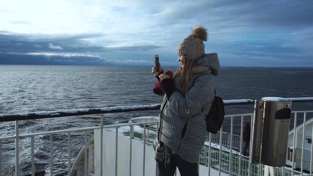 Adult woman in warm winter outwear and hat using smartphone and taking photo of frozen seascape while sailing on passage boat in sunlight.