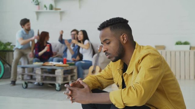 Young african amercan man feels upset and isolated while his friends celebrating party at home indoors