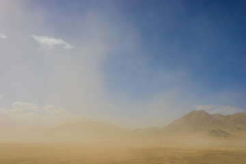 Wind blown sand drifts through the sky in front of rock and sand mountains in California desert.