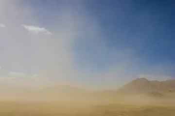 Gordijnen Wind blown sand drifts through the sky in front of rock and sand mountains in California desert. © kenkistler1