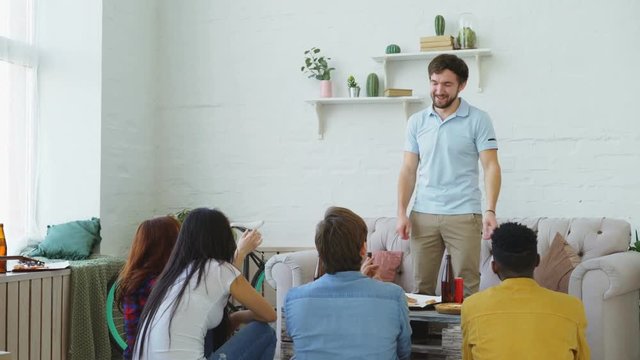 Group of friends having fun playing party game at home. Young guy showing something or somebody and his flatmates trying to guess it
