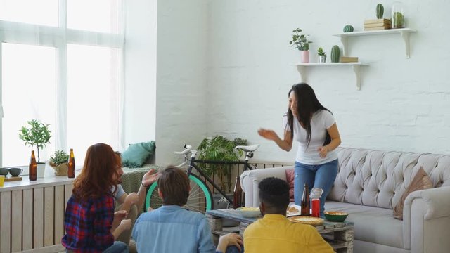 Group of multi-ethnic friends having fun playing party game at home. Young asian girl showing something or somebody and her flatmates trying to guess it