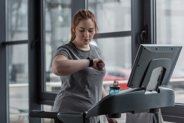 Overweight girl looking at fitness tracker while running on treadmill in gym