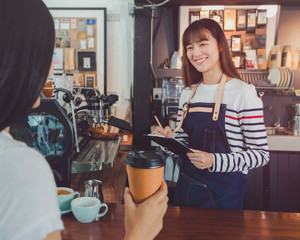 Young beautiful Asian woman barista wear blue apron talking to customer at bar counter in coffee shop with smile face.Concept of cafe and coffee shop small