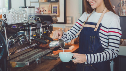 Young beautiful Asian woman barista wear blue apron holding hot coffee cup served to customer at bar counter in coffee shop with smile face.Concept of cafe and coffee shop small business.Vintage tone