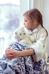 Cute little girl hugging a teddy bear. A cute baby in the room sits at the window in the winter