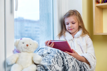 A cute baby in the room sits at the window in the winter and reads a book