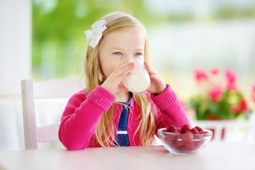 Pretty little girl eating raspberries and drinking milk at home. Cute child enjoying her healthy fresh organic fruits and berries.