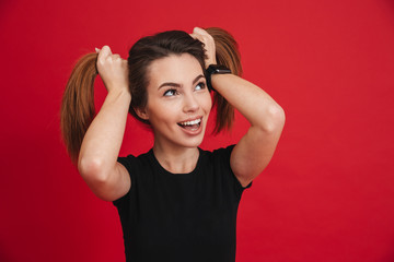 Cheerful childish woman 20s in black t-shirt having fun and making two ponytails with her brown hair, isolated over red background