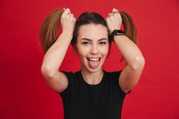 Image of amusing adult girl 20s in black t-shirt playing around and making two ponytails hairstyle with funny face, isolated over red background