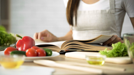 Housewife reading cooking book with fresh vegetables and kitchen tools on table