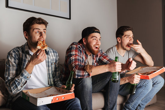 Image of three young bachelors eating pizza with pleasure, while watching football match at home tv