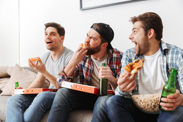 Portrait of three hungry men eating pizza and drinking beer with, while supporting football team at home