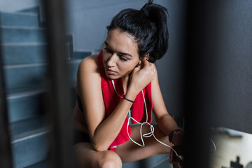 Naklejka premium Sporty attractive brunette female with red towel on neck after workout in the gym sits on the stairs and listening the music on earphones from smartphone. Sport concept.