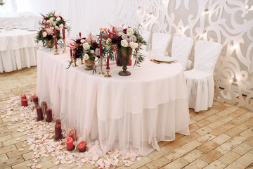 Decorate table with white tablecloth, red bouquets with fern and eucalyptus in brass vases, rose petals on the floor