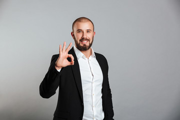 Image of young masculine man in business suit looking on camera and showing ok sign, isolated over gray background