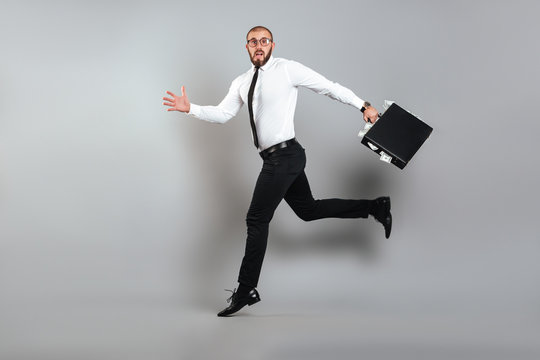 Image of young confused man in glasses and business suit running away with briefcase full of dollar bills in hand, isolated over gray background